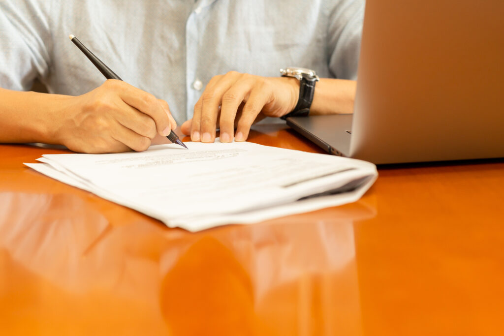 Businessman signsing documents with quill pen and laptop on wooden desk.